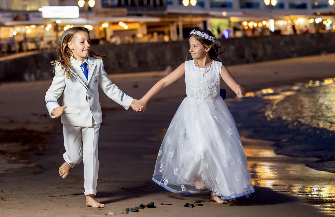 Dos niños vestidos de comunión corriendo de la mano por la orilla al anochecer
