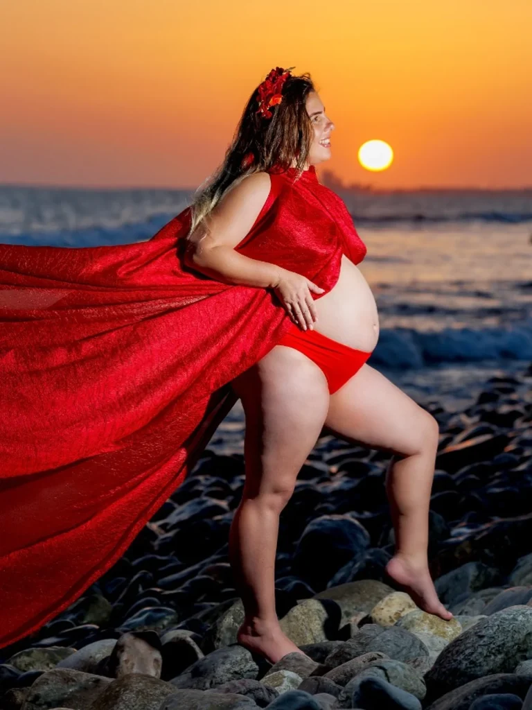 Embarazada con vestido rojo en una costa rocosa al atardecer