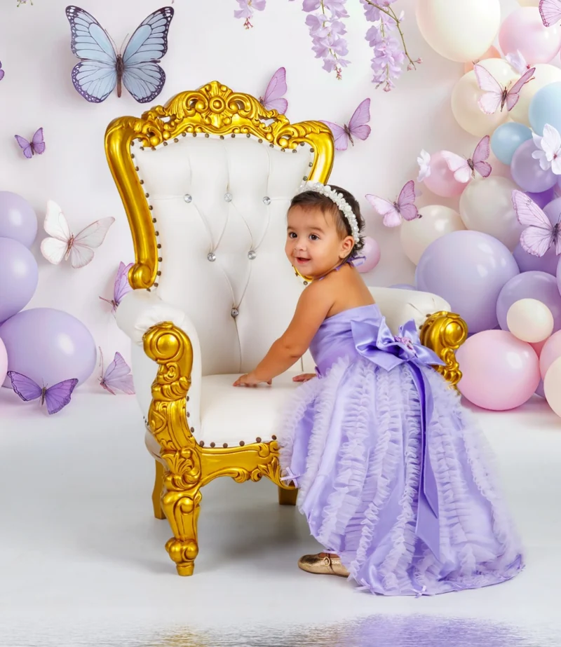 Niña con vestido lila sentada en un sillón blanco entre globos y mariposas